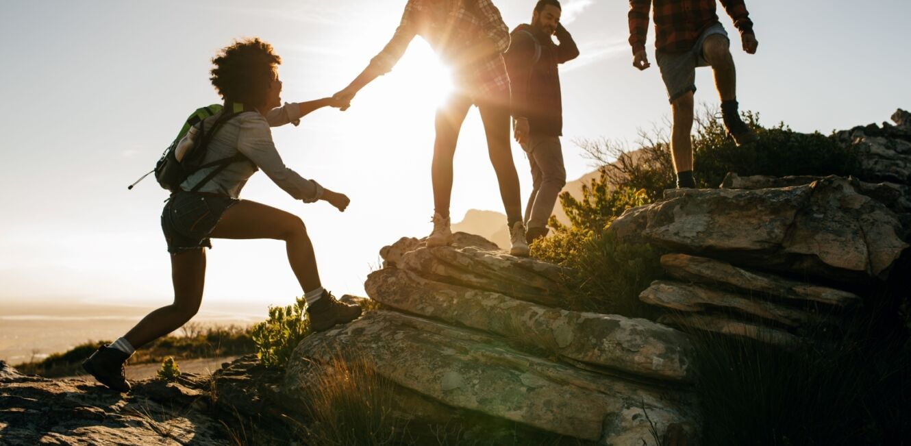 Gruppe von Wanderern hilft einander beim Aufstieg über felsiges Gelände bei Sonnenuntergang. Inklusion. Teamwork,